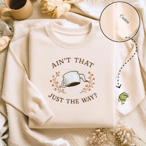 May include: Cream-colored sweatshirt with the embroidered phrase "Ain't That Just The Way?" and a teapot design. A small frog graphic is on the sleeve. The word "Custom" is printed on the sleeve.