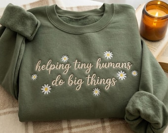 Op de afbeelding: Een olijfgroene sweatshirt met de geborduurde tekst "helping tiny humans do big things" in een sierlijk lettertype, versierd met kleine madeliefjes. De sweatshirt is op een patroon deken gevouwen, met een brandende kaars en een boek op de achtergrond.
