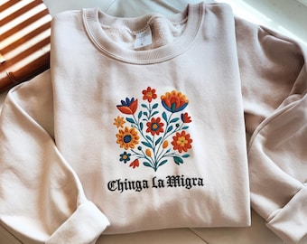 Puede incluir: Sudadera beige claro con un diseño de bordado floral en naranja, azul y amarillo. La frase "Chinga La Migra" está bordada en escritura negra debajo de las flores. Las mangas están dobladas.