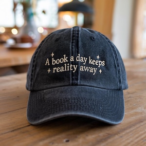 May include: A black baseball cap with the text "A book a day keeps reality away" embroidered in white on the front.