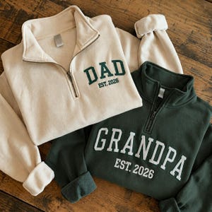 May include: A cream-colored quarter-zip sweatshirt with dark green embroidery reading DAD EST. 2026 and a dark green quarter-zip sweatshirt with white embroidery reading GRANDPA EST. 2026. Both garments feature a high collar and metal zipper.