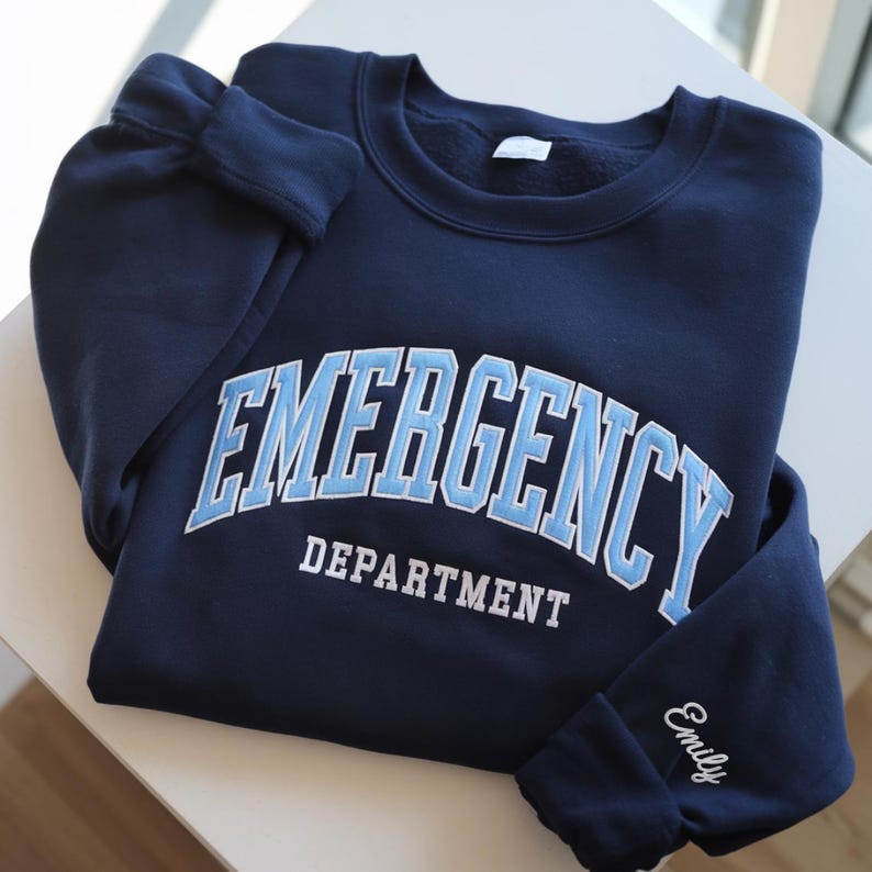 Op de afbeelding: Een marineblauwe sweatshirt met de tekst "EMERGENCY DEPARTMENT" in lichtblauw en wit op de borst. De naam "Emily" is in het wit op de mouw geborduurd. De sweatshirt is op een wit oppervlak gevouwen.