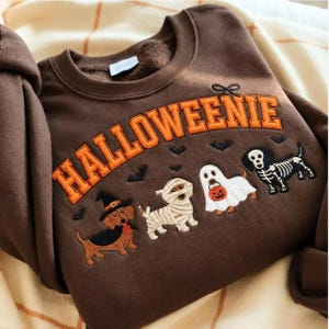 May include: A brown crewneck sweatshirt featuring the embroidered text HALLOWEENIE in orange letters. Below the text are four dachshunds dressed in Halloween costumes: a witch, a mummy, a ghost holding a pumpkin bucket, and a skeleton.