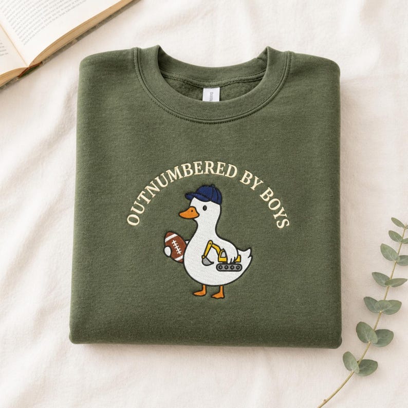 May include: Olive green sweatshirt with the text "OUTNUMBERED BY BOYS". The sweatshirt features a cartoon duck wearing a baseball cap and holding a football and a construction vehicle.