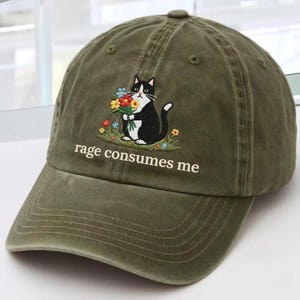 May include: Olive green baseball cap with an embroidered design of a black and white cat holding flowers. The text "rage consumes me" is embroidered below the cat. The cap has a curved brim and a fabric strap closure.