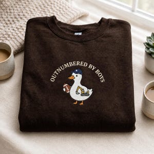 May include: A dark brown sweatshirt with the embroidered text "OUTNUMBERED BY BOYS". The design features a white duck wearing a blue cap and holding a football. The duck also has a small excavator on its belly.