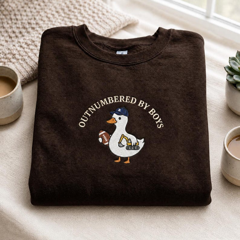 May include: A dark brown sweatshirt with the embroidered text "OUTNUMBERED BY BOYS". The design features a white duck wearing a blue cap and holding a football. The duck also has a small excavator on its belly.