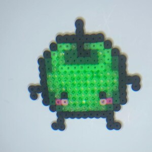 May include: A pixel art creation of a green alien-like character made from small, colorful beads. The figure has a square body, black outlines, and small arms and legs. The character has pink cheeks and a happy expression.