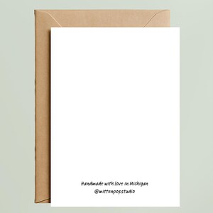 May include: A white greeting card with a brown envelope behind it. The card has the text "Handmade with love in Michigan @mittenpopstudio" in black script at the bottom. The background is a light green color.