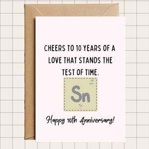 May include: A light pink anniversary card with a brown envelope. The card reads "Cheers to 10 years of a love that stands the test of time." and "Happy 10th Anniversary!" A graphic of the element Tin is in the center.