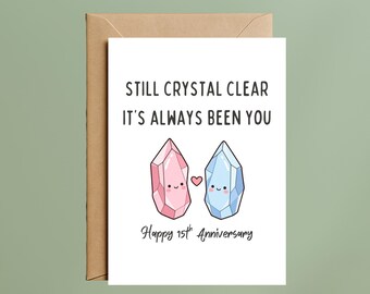 May include: A white greeting card with a brown envelope featuring a pink crystal and a blue crystal illustration with smiling faces and a small heart between them. The text reads: STILL CRYSTAL CLEAR IT'S ALWAYS BEEN YOU Happy 15th Anniversary.