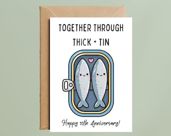 May include: A white greeting card with a brown envelope featuring a cartoon illustration of two grey fish inside a blue tin can. The text reads TOGETHER THROUGH THICK + TIN and Happy 10th Anniversary! The fish have smiling faces and pink cheeks with a small pink heart between them.