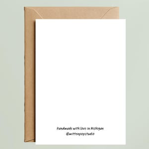 May include: A white rectangular greeting card with a brown kraft paper envelope. The bottom center of the card features black text that reads: Handmade with love in Michigan @mittenpopstudio. The card is blank and oriented vertically.
