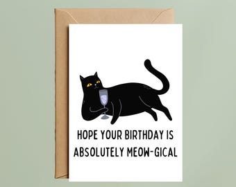 May include: A white greeting card featuring a black cat with yellow eyes holding a glass of champagne. The text below the cat reads: HOPE YOUR BIRTHDAY IS ABSOLUTELY MEOW-GICAL. The card is placed in front of a brown envelope.