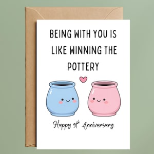 May include: A white greeting card with a brown envelope featuring the text Being with you is like winning the pottery, a small pink heart, two smiling pots in light blue and pink, and the text Happy 9th Anniversary at the bottom.