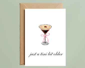 May include: A white greeting card featuring a watercolor illustration of an espresso martini in a stemmed glass with a pink ribbon tied around the stem and the text just a tini bit older printed in black script font below the cocktail.