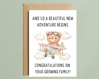 May include: A greeting card with a brown envelope. The card features a cartoon bear wearing goggles, flying a biplane with a pink propeller. Text reads, "AND SO A BEAUTIFUL NEW ADVENTURE BEGINS" and "CONGRATULATIONS ON YOUR GROWING FAMILY!"