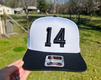 Può includere: Un cappellino da baseball bianco e nero con il numero 14 ricamato in nero sul davanti. Il cappellino ha una visiera piatta e una vestibilità classica. Il cappellino è tenuto da una mano.