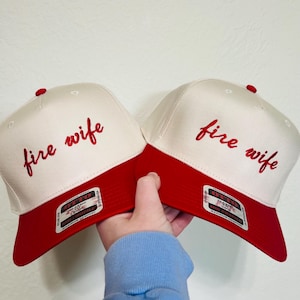 以下が含まれることがあります： 白いつばと赤いクラウンが付いた2つの野球帽。各帽子の前面には、赤いスクリプトで「fire wife」というフレーズが刺繍されています。各帽子のてっぺんには赤いボタンが付いています。