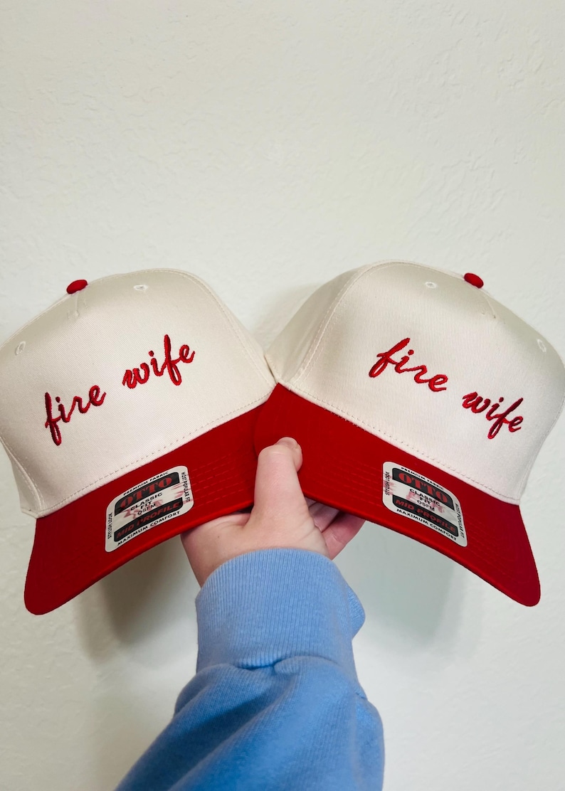 May include: Two baseball caps with a white crown and a red brim. The phrase "fire wife" is embroidered in red script on the front of each cap. A red button is on top of each cap.