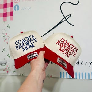 May include: Two baseball caps with a red brim and a cream-colored crown are held up. The text "COACH'S FAVORITE MOM" is embroidered on the front in navy blue and red. The caps have a red button on top.