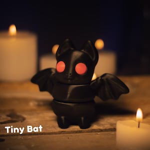May include: A black bat figurine with red eyes and outstretched wings. The text "Tiny Bat" is at the bottom. The bat is on a wooden surface, with lit candles in the background, creating a spooky atmosphere.