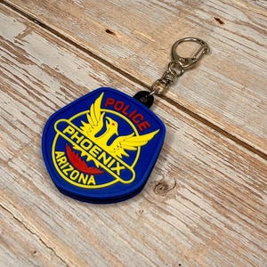 May include: A blue and yellow Phoenix Arizona Police badge keychain. The badge features the words "POLICE", "PHOENIX", and "ARIZONA". The keychain has a silver metal ring.