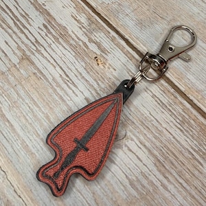 May include: A keychain featuring a textured, rust-orange arrowhead-shaped charm with a dark gray sword silhouette embroidered in the center. The charm is outlined in black stitching and attached to a silver-toned metal lobster claw clasp.