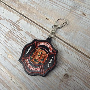 May include: Black and red Phoenix Fire Dept. keychain. The keychain features a shield design with the text "PHOENIX FIRE DEPT. EST. 1886" in white and red lettering. The keychain is attached to a silver-colored metal clasp.