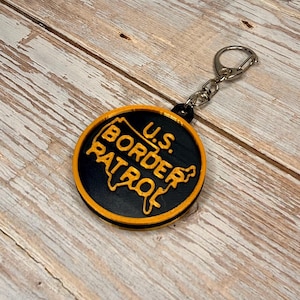 May include: A black and orange keychain with the text "U.S. Border Patrol" in a circular design. The keychain has a silver keyring attached. The design is on a wooden surface.