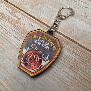 May include: A keychain featuring a Fire Department City of New York emblem. The shield-shaped patch has a red and gold border, with the text "FIRE DEPARTMENT CITY OF NEW YORK" in white. The emblem includes a stylized flame and building silhouettes.