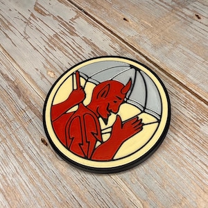 May include: A round, 3D-printed coaster featuring a red devil character holding an umbrella. The devil is set against a cream-colored background within a black-edged circle. The umbrella is gray.
