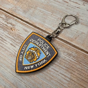 May include: A keychain featuring a shield-shaped emblem of the New York City Police Department. The emblem is blue, gold, and black, with the words "Police Department" and "City of New York" displayed. The keychain has a silver clasp.