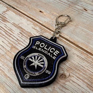 May include: Black and blue Police Glendale Arizona keychain. The keychain features a shield-shaped design with the words "POLICE GLENDALE" and "ARIZONA 1910" embroidered in white. A silver-colored metal clasp is attached.
