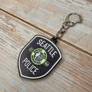 May include: A black and white Seattle Police badge keychain. The badge features the words "Seattle Police" and "Service, Pride, Dedication" around a central emblem. The keychain has a silver clasp and chain.