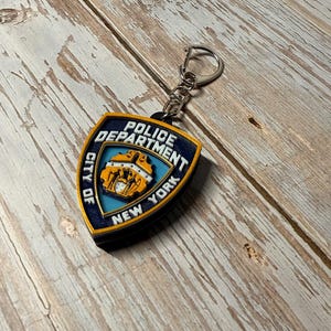 May include: A keychain featuring a shield-shaped emblem of the New York City Police Department. The emblem is in blue, gold, and black, with the words "Police Department" and "City of New York" displayed. The keychain has a silver keyring.