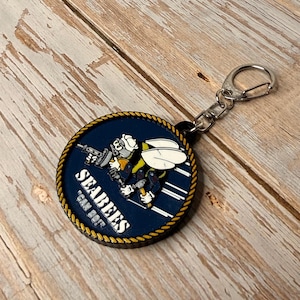 May include: A navy blue and gold round keychain featuring a cartoon bee holding a weapon, with the text "SEABEES" and "CAN DO!". The keychain has a silver-colored clasp and a rope-like border.