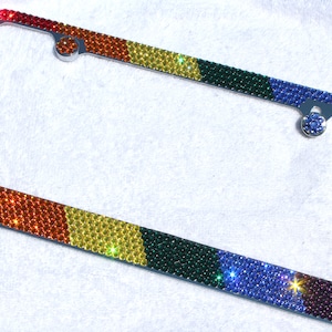 May include: A rainbow-colored license plate frame with rhinestones. The frame is made of metal and has a silver finish. The rhinestones are arranged in a pattern that resembles a rainbow.