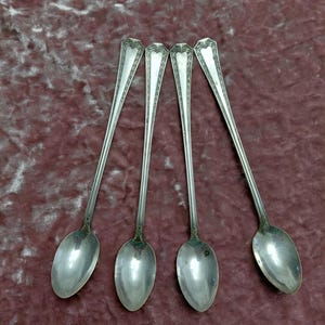 May include: A set of four long-handled silver-toned spoons featuring slender stems and decorative embossed detailing at the top of each handle. The spoons have oval-shaped bowls and a polished metallic finish, arranged side by side in a row.