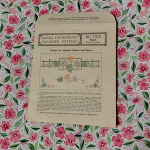 May include: A vintage McCall Kaumagraph transfer pattern for appliqué bolster and sprays. The pattern is on a cream-colored paper with a floral design. The text includes "No. 1570 BLUE" and "Price 30 cents". The background is a floral fabric.