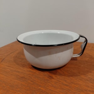 May include: A white enamel chamber pot with a black rim and a single side handle. The bowl has a rounded shape and a smooth finish, featuring a wide opening at the top. The exterior surface is plain white with a black trim along the edge of the handle.