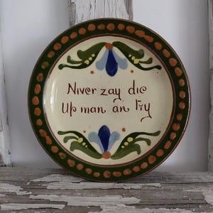 May include: A round ceramic plate with a cream-colored center featuring the hand-painted text Niver zay die Up man an Fry in brown script. The plate is decorated with blue and green floral motifs and a dark green rim with orange dots.