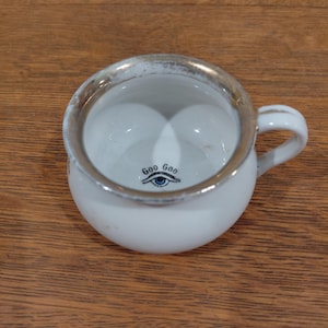 May include: A small white ceramic chamber pot with a single handle and a metallic silver rim. The interior base features a graphic of a blue eye with the text "Goo Goo" printed above it in black lettering, centered at the bottom of the basin.