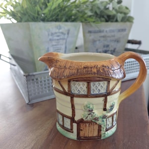 May include: A ceramic creamer pitcher shaped like a cottage with a thatched roof, timber-framed walls, and a small wooden door. The pitcher features a cream-colored base, brown roof details, and a curved handle, with a spout for pouring liquids.