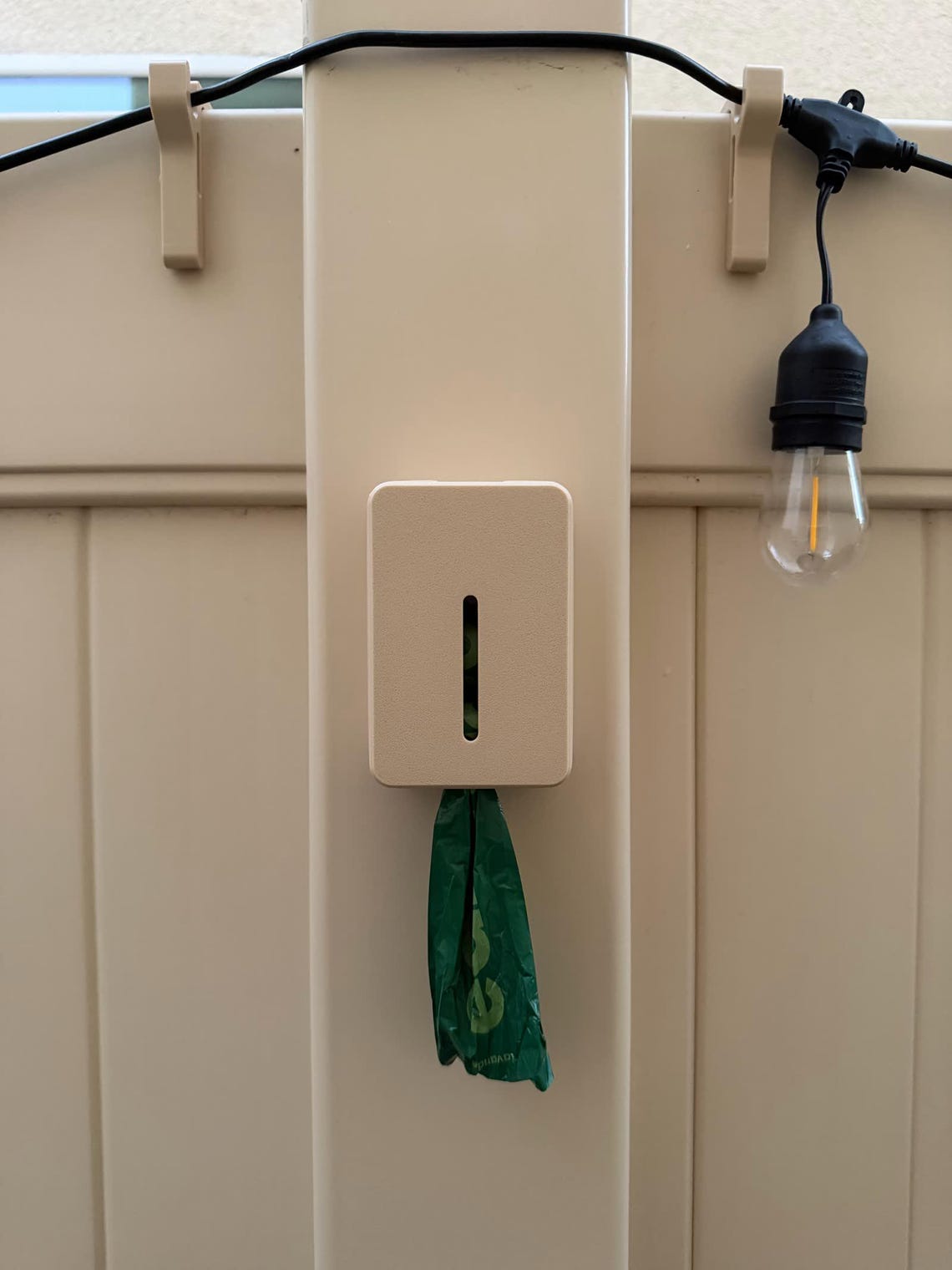 Beige poop bag dispenser on a beige vinyl fence post, a green bag hanging from the bottom and string lights on the rail above