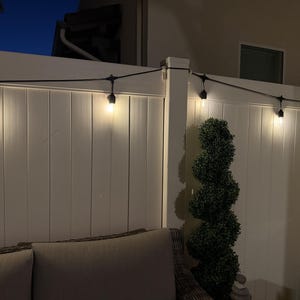 May include: A string of outdoor Edison-style light bulbs with black sockets and wiring, emitting a warm yellow glow. The lights are suspended horizontally against a white vertical panel fence, creating a series of illuminated points.