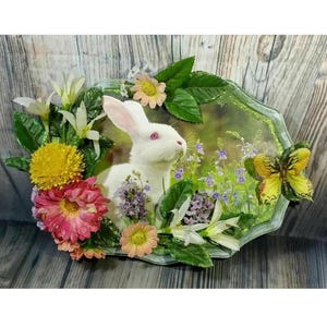 Könnte beinhalten: Ein weißes Kaninchen mit rosa Augen sitzt in einem Blumenfeld. Das Kaninchen ist von verschiedenen Blumen umgeben, darunter gelbe, rosa und lila. Ein Schmetterling sitzt auf einem Blatt in der oberen rechten Ecke des Bildes.