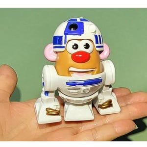 May include: A white and blue R2-D2 themed Mr. Potato Head toy. The toy has a white body with blue details and a round head with a white and blue helmet. The toy has a red nose and black eyes.