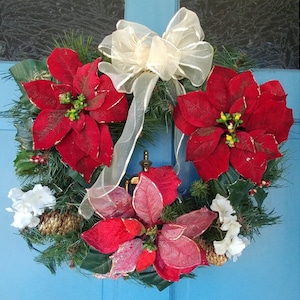 Puede incluir: Una corona de Navidad con flores de poinsettia rojas, ramas de pino verdes, flores blancas y un lazo dorado.