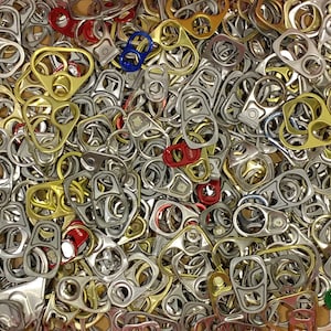 May include: A collection of assorted metal can pull tabs in various colors, including silver, gold, red, and blue. The tabs are densely packed together, creating a textured surface. The image is a close-up shot, highlighting the details of each tab.